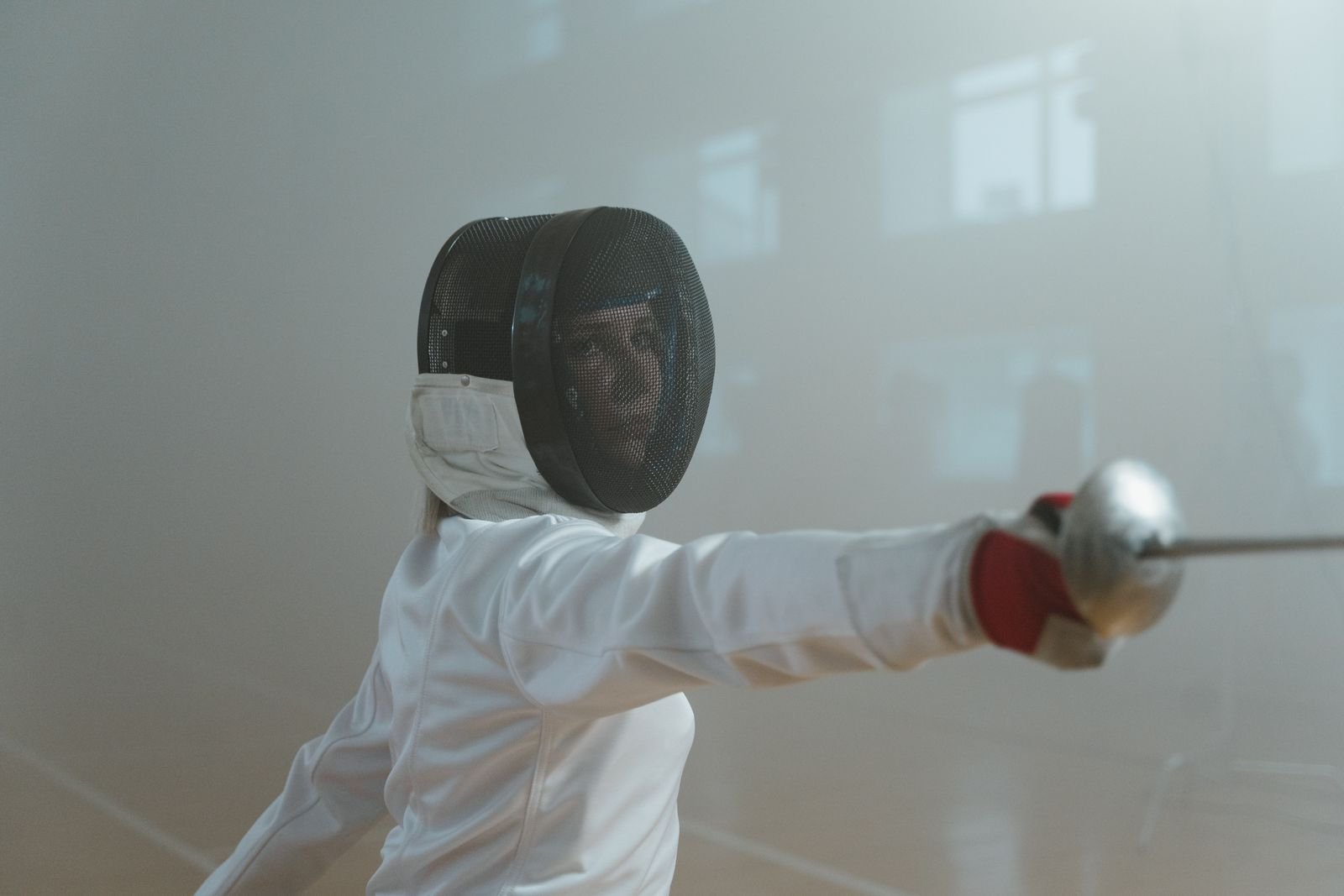 A masked fencer in a deep lunge extending the blade in a bright training hall.