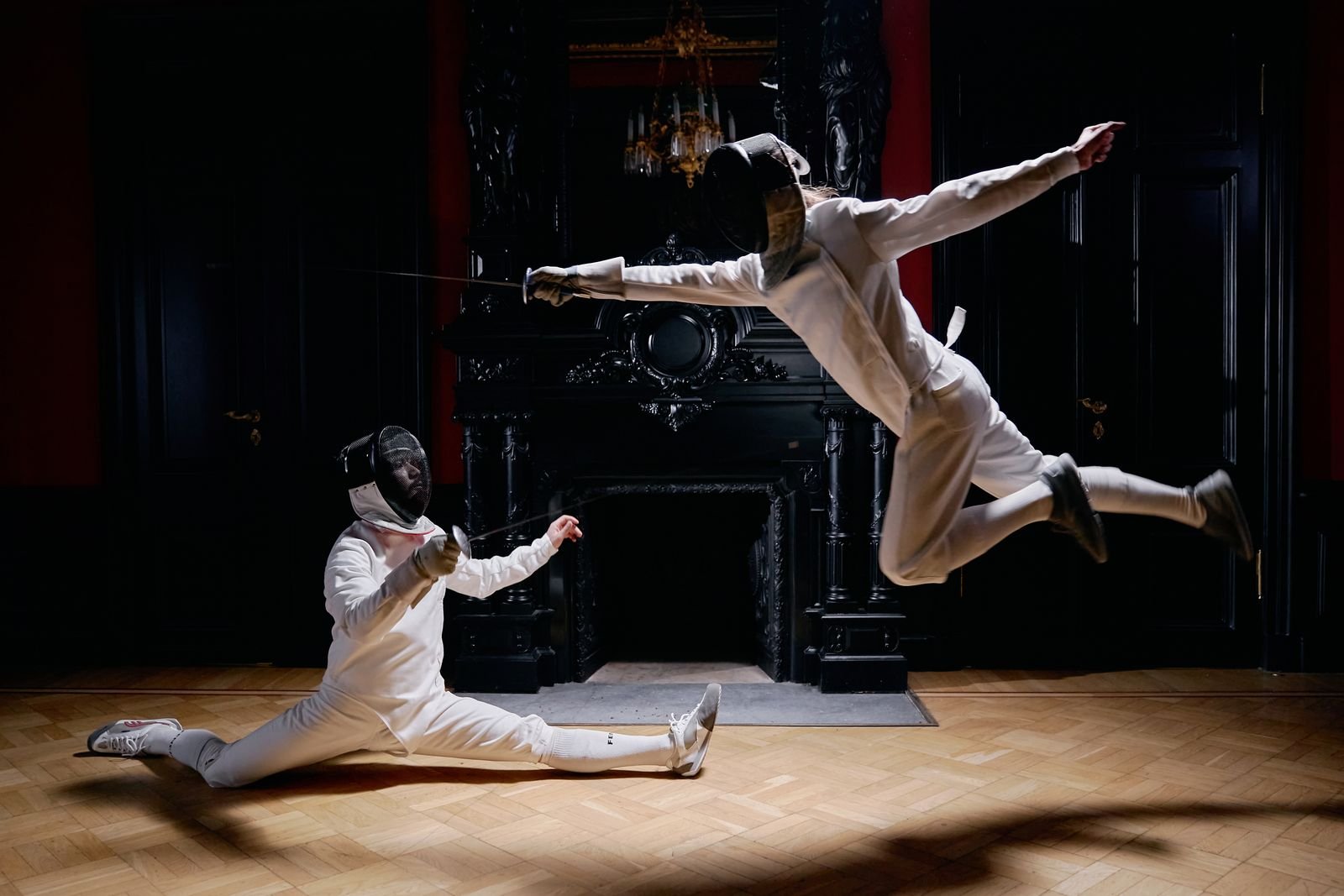 Two fencers in a dark, chandelier-lit hall — one in a deep lunge, one leaping mid-air, blades extended.
