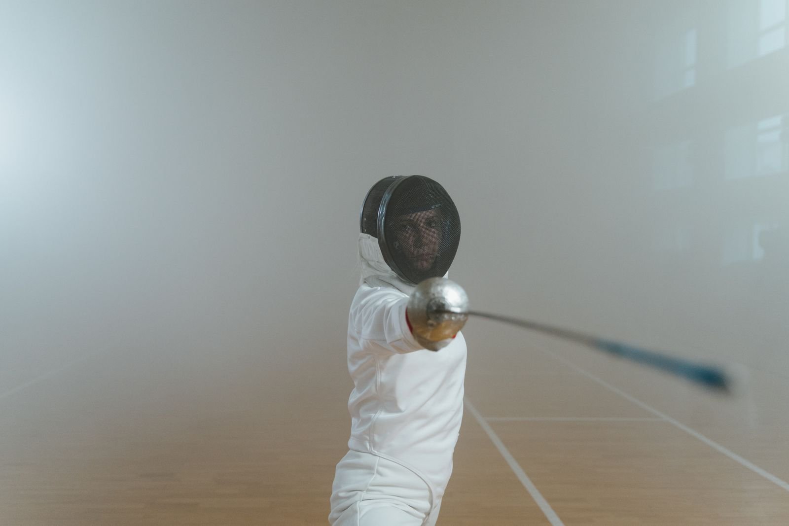 A single masked fencer holding a blade upright in a hazy hall.